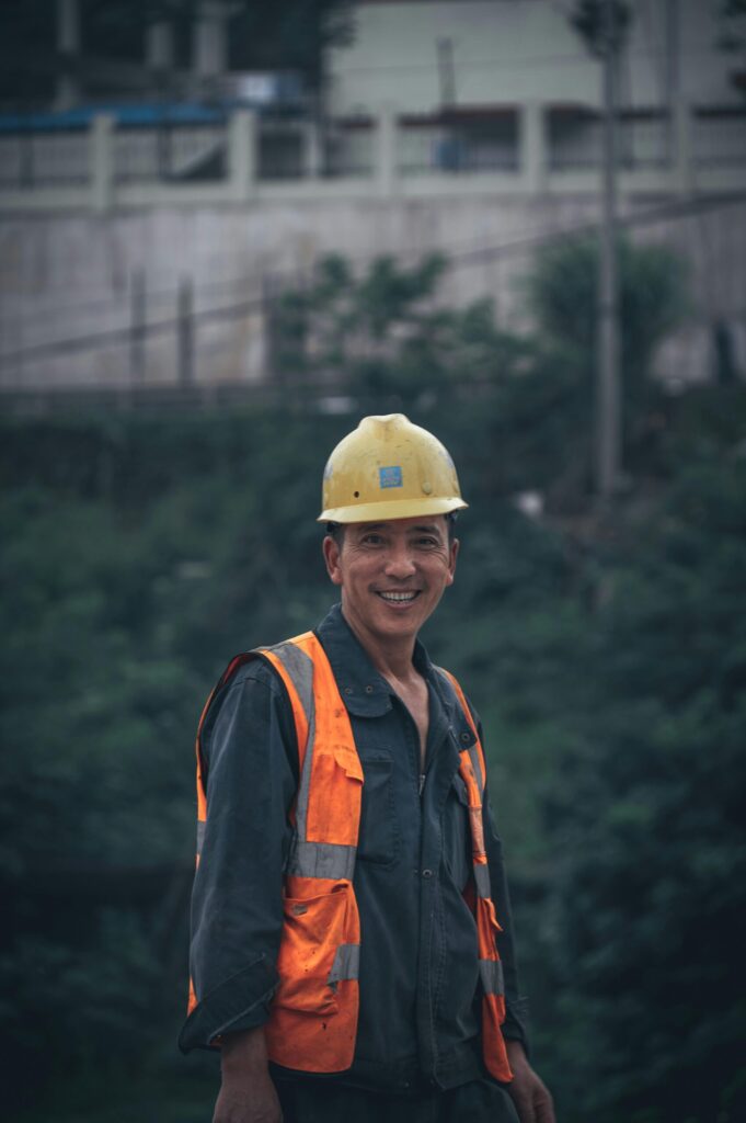 pexels-photo-12357629-12357629 Un obrero sonriente con equipo de seguridad está de pie al aire libre en un entorno de trabajo.