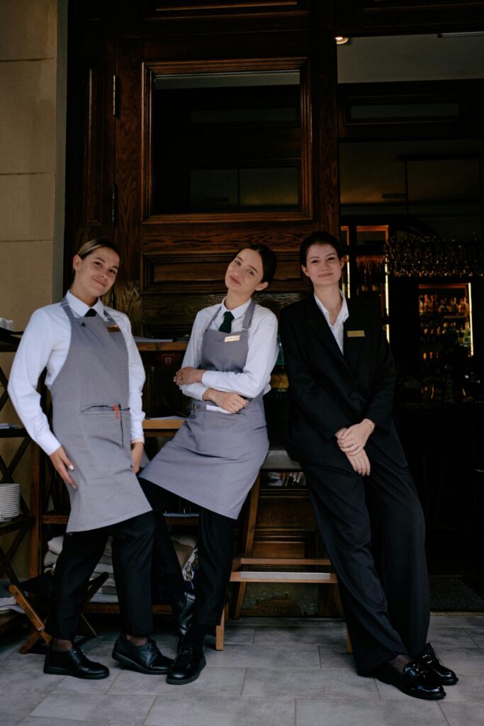 pexels-photo-33934080-33934080 Tres miembros del personal de un restaurante con uniformes formales posando alegremente frente a un restaurante.